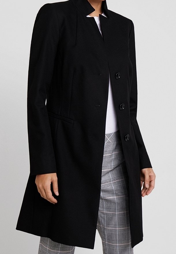 Manteau noir sur mesure avec un col haut, tissu texturé, trois boutons et poches latérales, associé à un pantalon à carreaux.