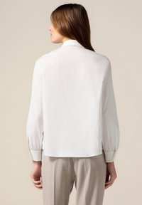 Chemise blanche à manches longues avec une coupe décontractée, en tissu doux, et un léger plissage aux poignets. Associée à un pantalon gris clair.