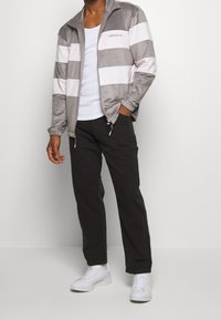 Ljust grå och vit randig zip-up jacka med hög krage, bärs över en vit tanktop och kombineras med mörka byxor och vita sneakers.