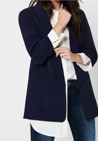 Marinblå kavaj med skräddarsydd design, två framfickor och långa ärmar. Bärs över en vit skjorta med uppvikta ärmar, i kombination med blå jeans.