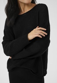 Black knitted sweater with a loose fit, wide neckline, and long sleeves. Features a textured pattern and raw edges at the hem and cuffs.