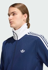 Tamsiai mėlynas užtrauktuku užsegamas švarkas su aukštu baltu apykaklės kraštu, trimis baltomis juostelėmis ant pečių ir balta logotipo siuvinėjimu krūtinėje. Lygus, tekstūruotas audinys.