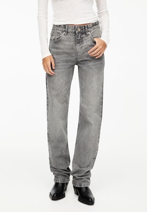 Personne portant un jean taille haute gris délavé à coupe droite, des bottes noires à bout carré et un haut blanc ajusté à manches longues, debout sur fond blanc.