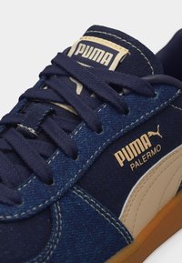 Detailný záber na námornícky modrú tenisku Puma Palermo s béžovým logom, detailmi šitia, námorníckymi šnúrkami a podrážkou gumovej farby.