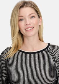 Uśmiechnięta kobieta z prostymi blond włosami w czarno-biało paskowanym swetrze, pozująca na jednolitym jasnym tle.