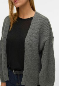 Szary sweter z dzianiny z otwartym przodem, luźnym krojem i prążkowaną fakturą, noszony na czarną bluzkę i zestawiony z ciemnymi dżinsami oraz czarnym paskiem.