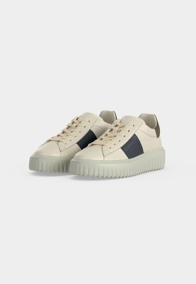 Paire de baskets écru avec semelles épaisses texturées, lacets beiges et bande latérale noire, présentée sur un fond clair uni.