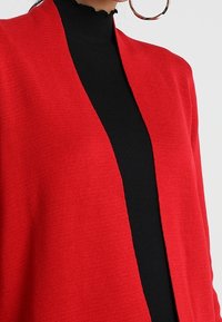 Cardigan rosso aperto davanti realizzato in tessuto a maglia texturizzata, indossato sopra un top nero a collo alto, caratterizzato da un design drappeggiato e privo di chiusure.