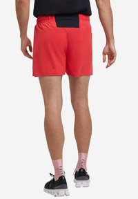 Pantaloni sportivi rossi con cintura nera; tessuto leggero; presenta un orlo a spacco; indossati con scarpe da corsa nere e calze rosa.