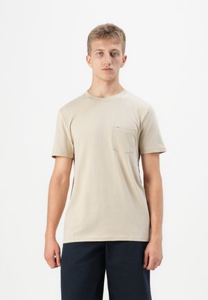 Ung man som bär en beige kortärmad t-shirt med bröstficka och mörka shorts, stående framför en enfärgad vit bakgrund.