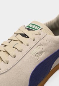 Κοντινό πλάνο μπεζ αθλητικού παπουτσιού Puma με κρέμ κορδόνια, πράσινο και μαύρο λογότυπο Puma στη γλώσσα, και μπλε ρίγα στο πλάι.