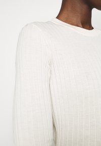 Světlý béžový žebrovaný pletený svetr s dlouhými rukávy a kulatým výstřihem. Strukturální vzor tvoří jemné svislé linie po celé tkanině.