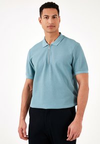 Gaiši zils polo krekls ar rāvējslēdzēja apkakli, īsām piedurknēm un teksturētu audumu, apvienots ar tumšiem biksēm. Vienkāršs dizains, nav acīmredzamu rakstu.