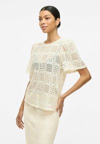 Top de color beige claro con mangas cortas, que presenta un patrón de cortes geométricos en toda su superficie, fabricado con un material transpirable.