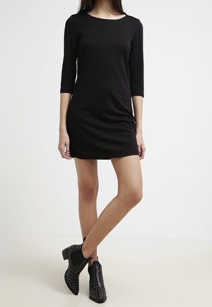 Woman wearing a black, three-quarter sleeve mini dress paired with black studded ankle boots, standing with one leg crossed over the other.