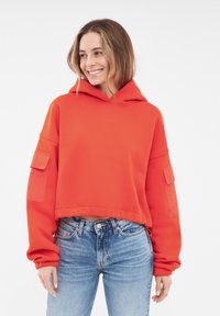 Rode cropped hoodie van zacht materiaal met een voorzak in kangoeroe-stijl en patchaccenten op de mouwen, gecombineerd met lichtblauwe high-waisted jeans.