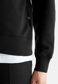 Juodas džemperis su struktūruota medžiaga, apatinėje dalyje matyti balta vidinė sluoksnis. Šone yra mažas logotipo žymeklis.