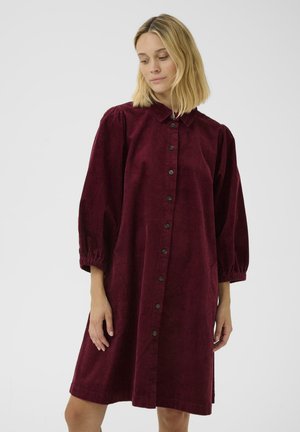 Vrouw in een bordeauxrode corduroy jurk met lange poffende mouwen en zwarte knopen aan de voorkant, die iets naar beneden kijkt.