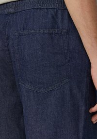 Pantalons en denim foncé avec une poche arrière, texture tissée et une coupe décontractée avec une taille élastique. Détails de couture classiques visibles.