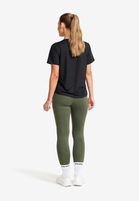 Kvinna med långt hästsvans iförd svart t-shirt, olivgröna leggings, vita strumpor och vita sneakers, stående vänd bort från kameran mot vit bakgrund.