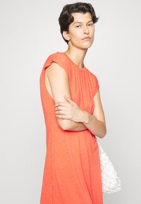 Vestido curto amarelo-avermelhado de manga curta com um corte solto, apresentando pregas nos ombros. Acompanhado por uma bolsa de rede branca. Fundo liso.