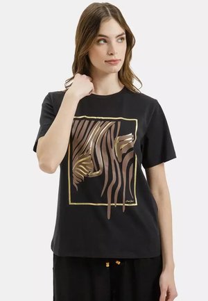 Kobieta w czarnej koszulce z metalicznym złotym i brązowym abstrakcyjnym wzorem pasków zebry w złotej kwadratowej ramce.