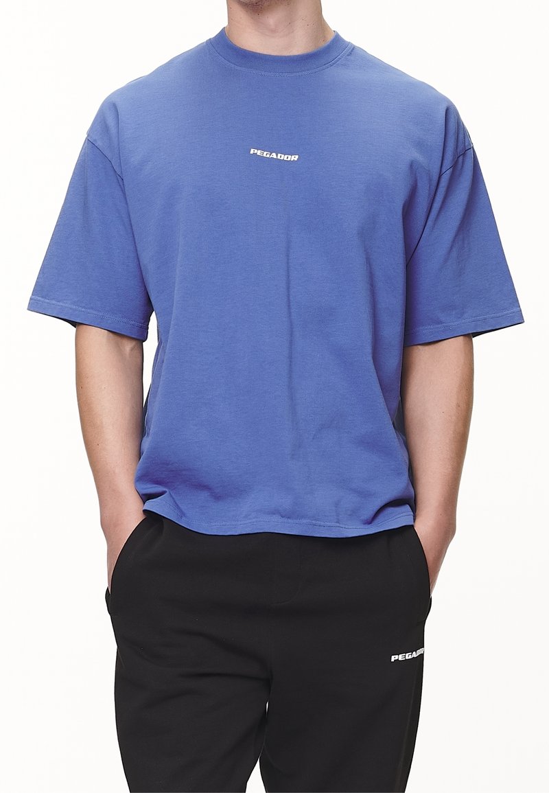 Personne portant un T-shirt bleu à manches courtes avec le texte "PEGADOR" et un pantalon noir avec le logo blanc "PEGADOR", mains dans les poches.
