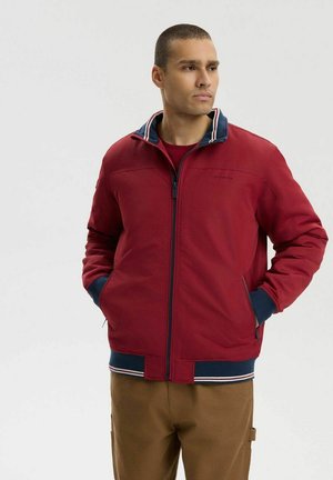 Mężczyzna w czerwonej kurtce z zamkiem błyskawicznym, granatowymi i białymi lamówkami, brązowych spodniach, stojący z rękami w kieszeniach, patrzący na bok na białym tle.