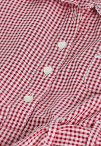 Punainen ja valkoinen ruudullinen paidankangas, jossa on näkyvät valkoiset napit ja yksityiskohtainen ompelu. Kankaan pinta on sileä ja klassinen gingham-kuvio.