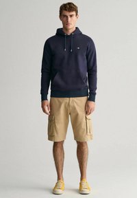 Marine sweatshirt med snørebånd, kængurulomme; beige cargo shorts med sidelommer; gule sneakers; modellen står foran en neutral baggrund.