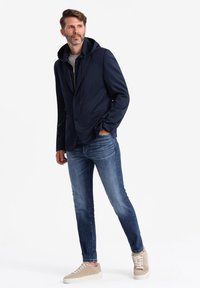Marineblazer med hætte, med jakkelommer, båret over en lys skjorte og blå jeans. Lyse sneakers fuldender outfittet.