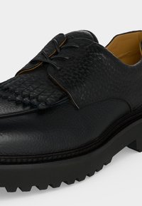 Chaussure noire en cuir à lacets avec dessus texturé, franges décoratives sur le cou-de-pied et semelle épaisse rainurée.
