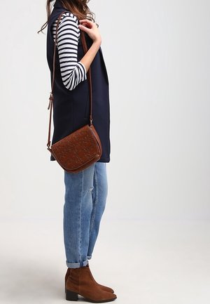 Mujer con camisa de rayas, chaleco azul marino, vaqueros azules, botines marrones y lleva un bolso cruzado de cuero marrón con relieve.