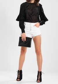 Blusa negra transparente con mangas con volantes, pantalones cortos blancos desgastados con botones al frente, bolso de mano negro y tacones negros con cordones.