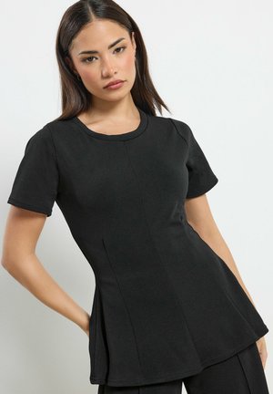 Femme aux cheveux raides et foncés portant un haut noir ajusté à manches courtes avec un col rond, posant devant un fond clair uni.