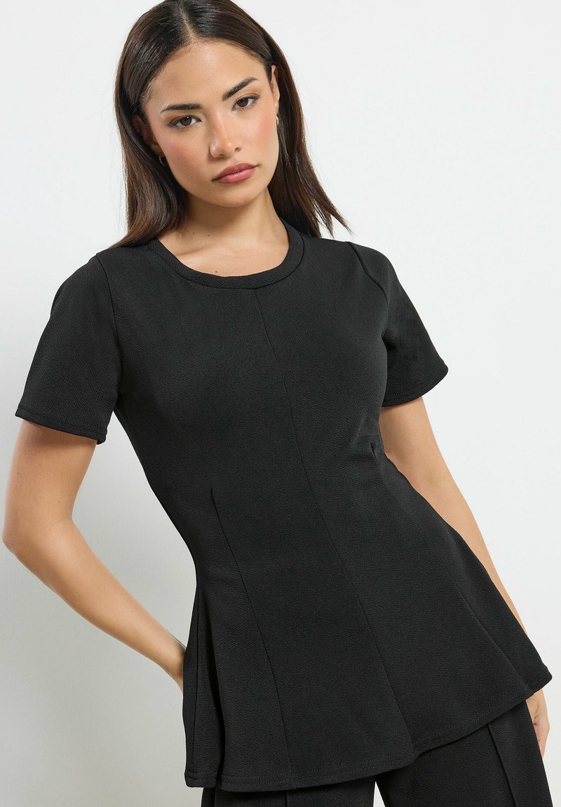 Femme aux cheveux raides et foncés portant un haut noir ajusté à manches courtes avec un col rond, posant devant un fond clair uni.