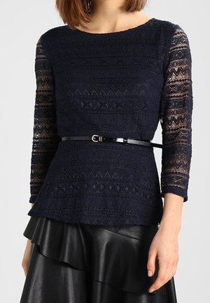 Nainen, joka pukeutunut tummansiniseen pitsiseen pitkähihnaiseen yläosaan mustan vyön kanssa ja mustaan nahkahameeseen, jossa on röyhelöinen helma, seisoo yksivärisen taustan edessä.