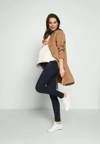 Casaco trench bege sobre uma t-shirt branca, jeans skinny escuros e ténis brancos. O conjunto apresenta um corte descontraído e um design casual.