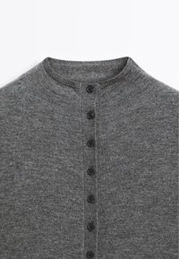 Pull en maille grise avec un col rond et sept boutons noirs sur le devant, fabriqué dans un tissu doux et texturé.