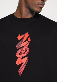 Primer plano de una persona que lleva una camiseta negra con un gráfico vertical en negrita de color rojo centrado en el pecho.