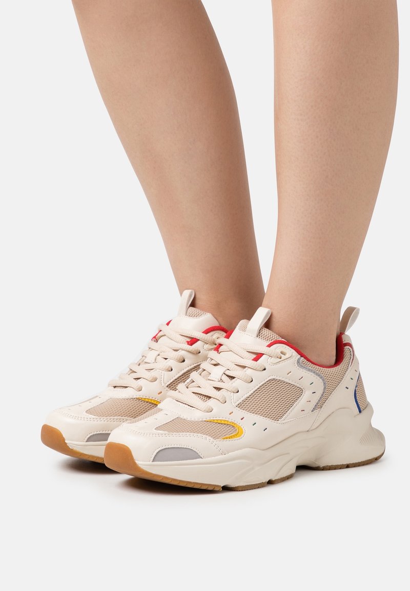Pieds portant des baskets beige épaisses avec des panneaux en maille, des bordures rouges et des semelles en caoutchouc sur un fond blanc uni.