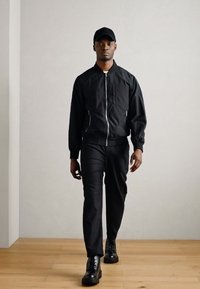 Schwarze Bomberjacke mit Reißverschluss und Seitentaschen, kombiniert mit schwarzen Hosen und klobigen schwarzen Stiefeln. Das Model trägt eine passende schwarze Mütze.