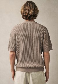 Suéter de punto beige con un patrón texturizado, mangas cortas y un dobladillo acanalado. La vista trasera resalta el corte holgado y los detalles de costura.