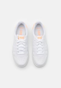 Par vita Asics-sneakers med ljusa persikofärgade detaljer, snörade och sedda uppifrån mot en enfärgad vit bakgrund.