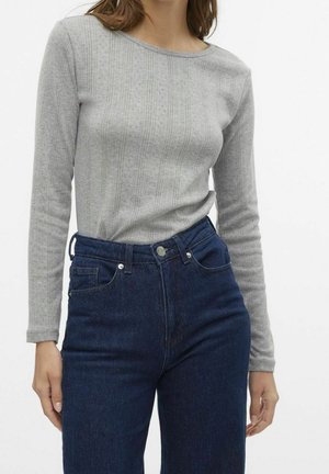 Frau trägt einen hellgrauen, langärmligen Strickpullover mit dezenten vertikalen Mustern, der in hoch taillierte dunkelblaue Jeans gesteckt ist.