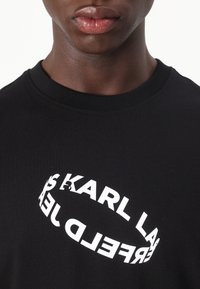 Melna kreklforma T-kreklis no kokvilnas, ar apļveida balto grafiku, kurā redzams teksts "KARL LAGERFELD" dažādās orientācijās.