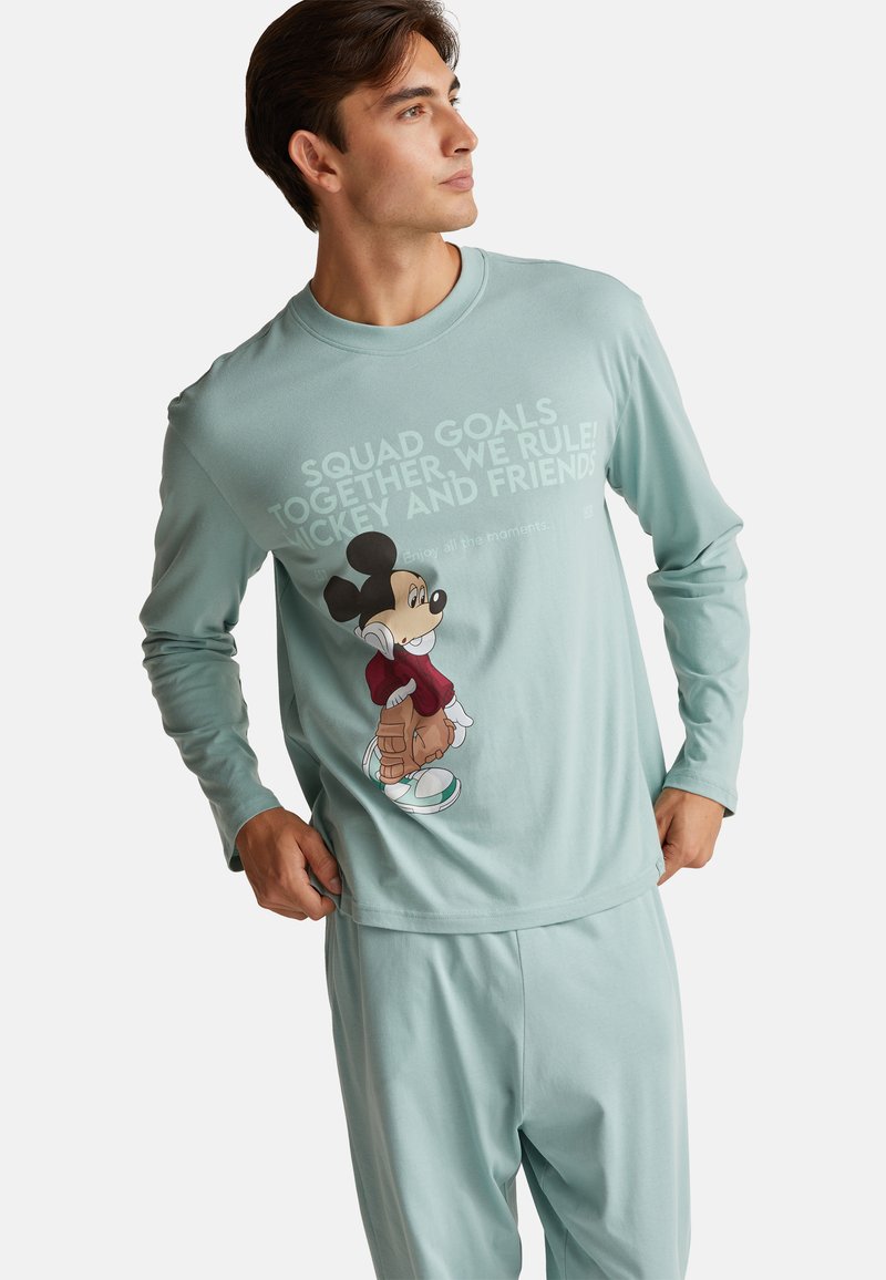 Mladý muž v bleděmodré košili s dlouhým rukávem a kalhotách s grafikou Mickey Mouse a sloganem o přátelství, stojící s rukama u kapes.