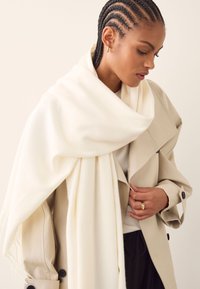 White scarf draped over a cream coat, featuring a soft texture. The outfit includes a black bottom and a layered design with visible buttons.