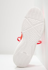 Bílá gumová podrážka tenisky Adidas s texturovaným vzorem. Jasně červené akcenty a odpovídající tkaničky, které ukazují na elegantní design.