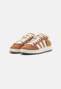 Sneaker in suede marrone con strisce e lacci bianchi, dettagli beige, punta rotonda e suola testurizzata. Design classico low-top.